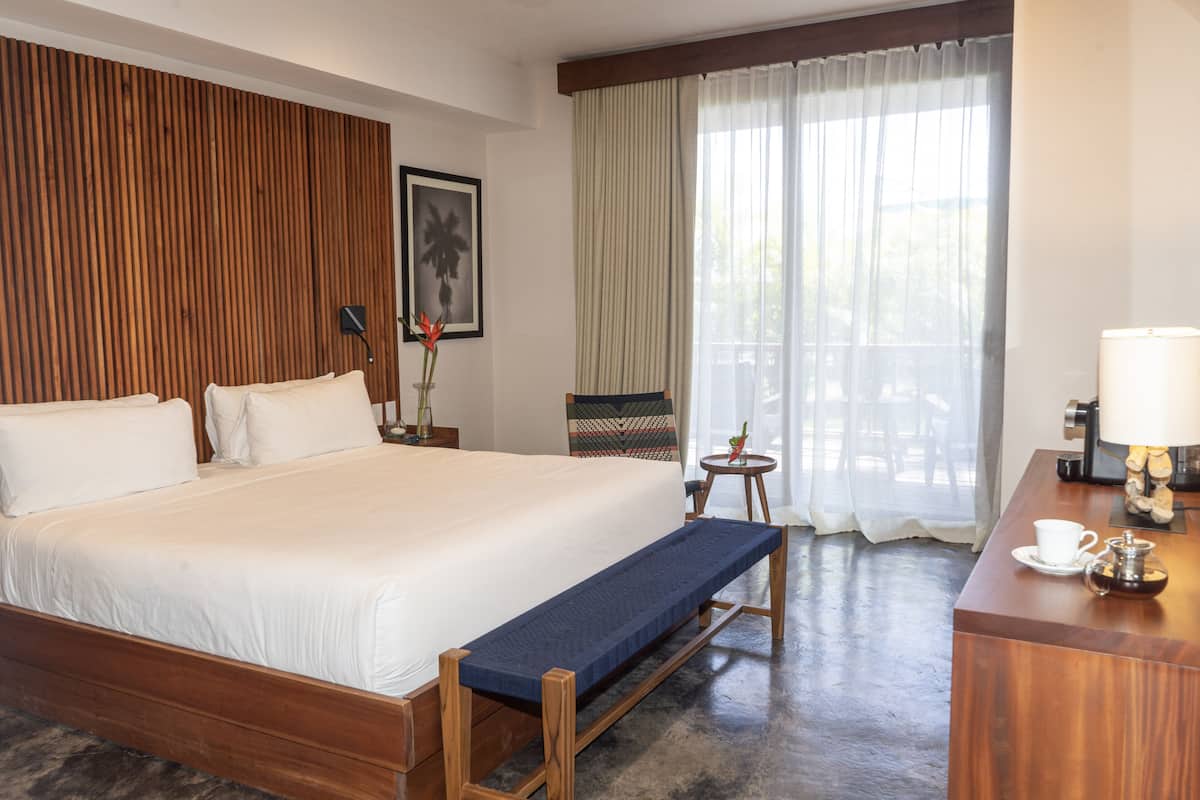 A hotel room with a large bed, a bench, a desk with a lamp, a coffee maker, and a view of the outdoors through sheer curtains.