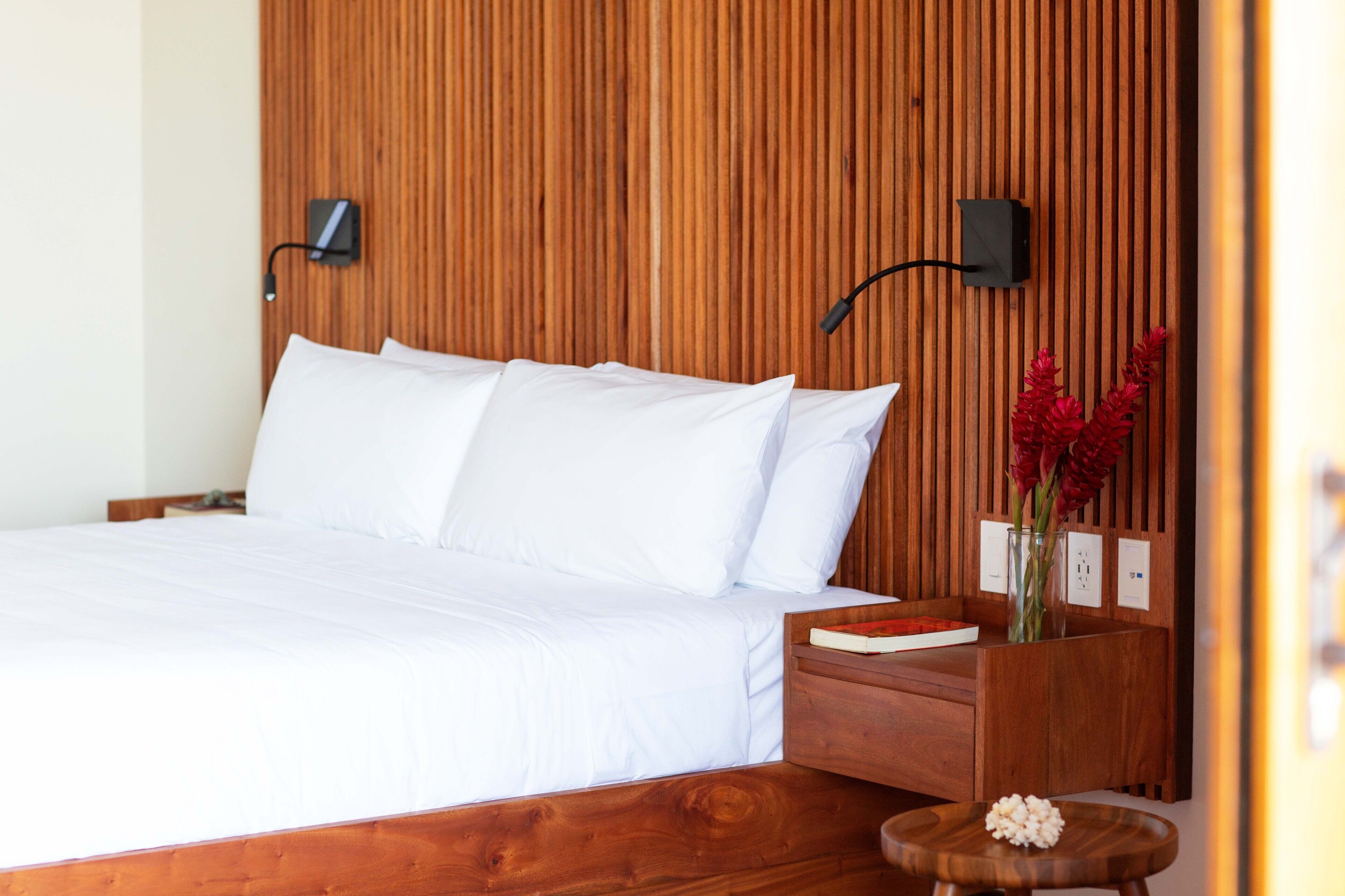 A neatly made bed with white linens, a wooden headboard, a bedside table with a book and a vase of flowers, and a wall-mounted lamp.