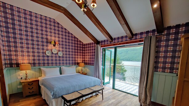 Un dormitorio con pared a cuadros, cama con cubrecama azul, suelo de madera y vista a un lago a través de un ventanal.