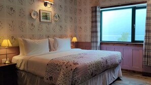 Une chambre avec un grand lit, deux lampes de chevet, un papier peint à motifs floraux, une fenêtre avec des rideaux et une vue sur l’extérieur.