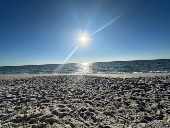 Una playa con huellas que conducen al agua, cielo despejado y el sol brillando.
