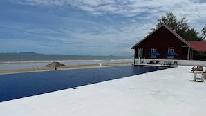 Een zwembad met uitzicht op het strand en de oceaan, een rood gebouw met blauwe parasols, en een rieten constructie in het zand.