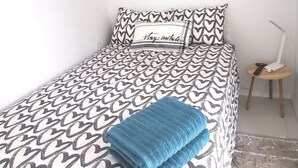 Ein ordentlich bezogenes Bett mit einer zudeckdecke in Herzmuster, ein blaues Handtuch auf dem Bett und ein kleiner Nachttisch mit einer Lampe daneben.