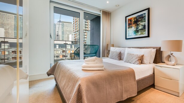 A modern hotel room with a large bed, a bedside table, a nightstand, a framed picture on the wall, and a view of the city through the window.