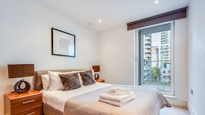 A modern bedroom with a large bed, a bedside table with a lamp, a framed artwork on the wall, and a view of high-rise buildings through a large window.