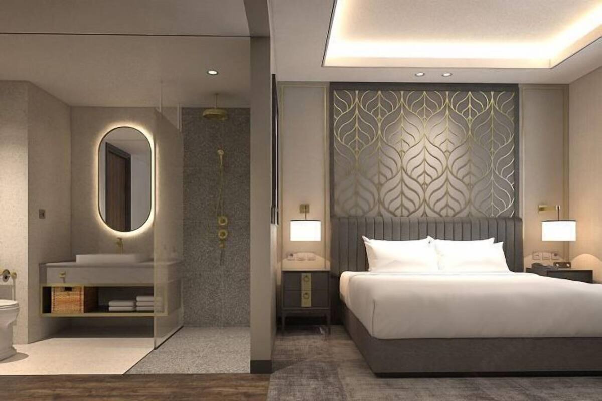 A modern hotel room with a large bed, a bedside table with a lamp, a vanity with a mirror, and a decorative wall panel.