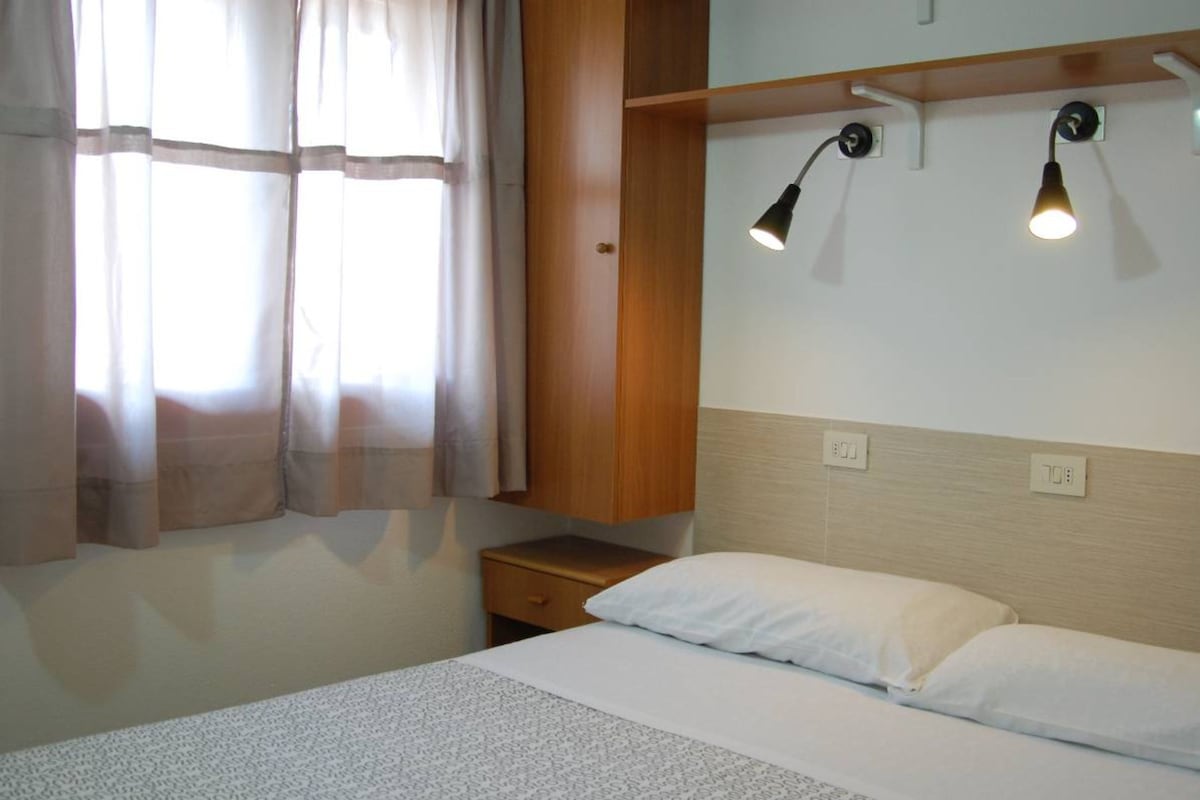 A hotel room with a bed, a nightstand, a wooden shelf, and two wall-mounted reading lamps.