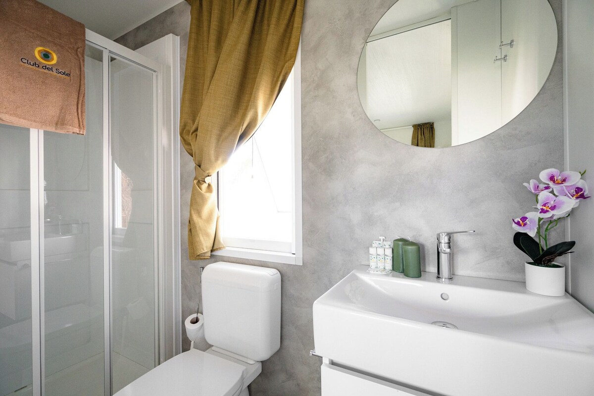 A modern bathroom with a glass shower enclosure, a toilet, a sink with a mirror, and a window with curtains.