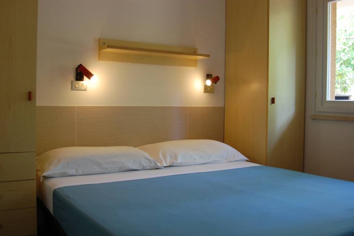 A bedroom with a bed, two bedside lamps, a wooden headboard, and a window.