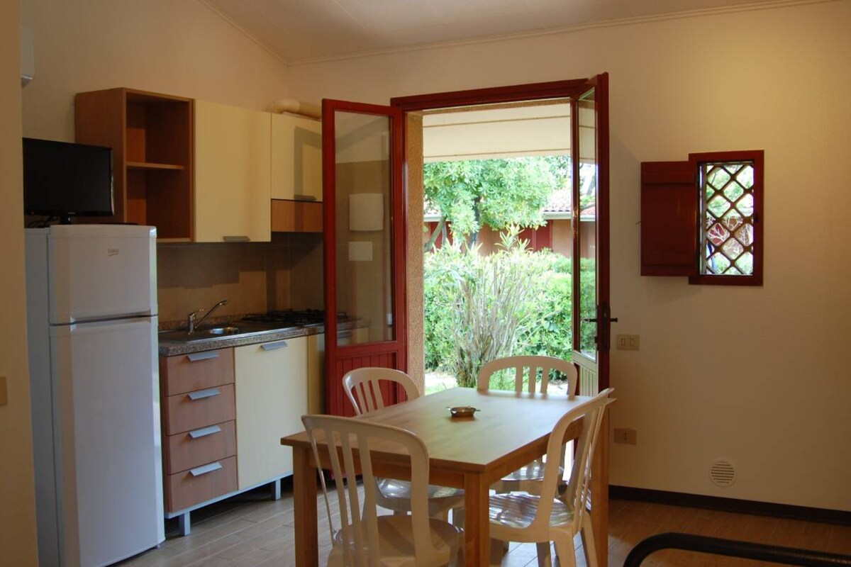 A compact kitchen with a refrigerator, stove, and wooden cabinets. There is a dining table with chairs and a window with a view of greenery outside.