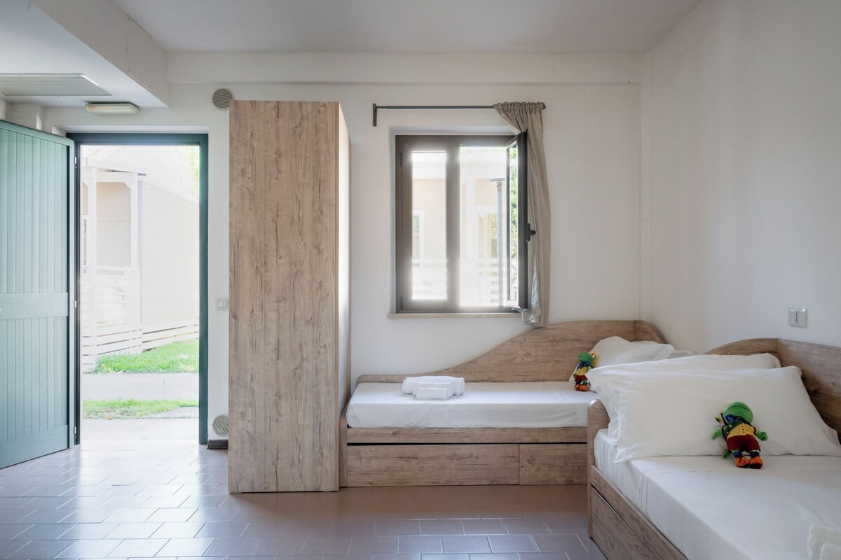 A bedroom with a bed, a window with curtains, a wooden door, and a view of the outside.