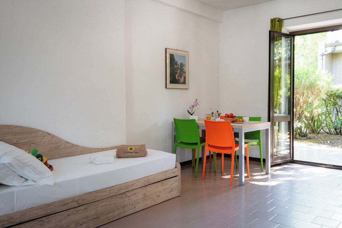 A bedroom with a bed, a small table, and chairs. There is a window with a view of greenery outside.