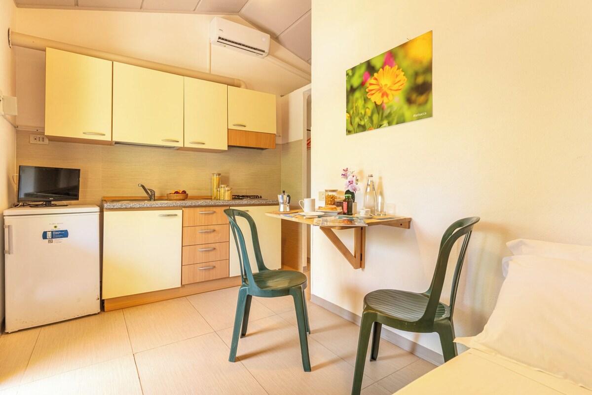 A compact kitchen with a refrigerator, stove, and sink. There are two chairs and a small table with a picture on the wall.