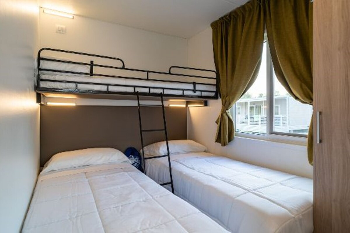 A dormitory room with bunk beds, a window with curtains, and a bed with a pillow and a blanket.