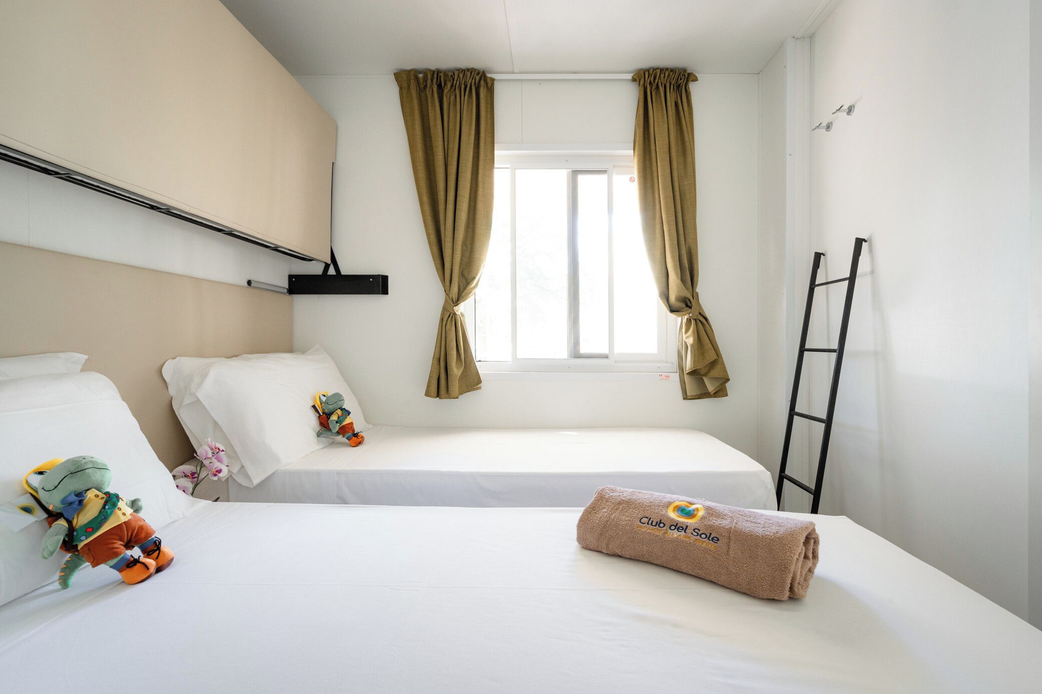 Ein Hotelzimmer mit zwei Betten, einem Fenster mit Vorhängen, einem Handtuch mit Logo und einer an der Wand stehenden Leiter.