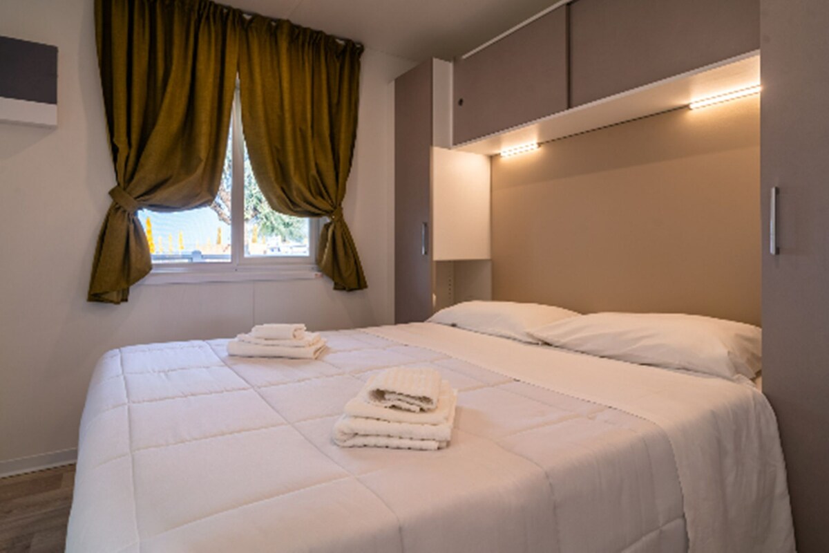 A neatly made bed with white linens and two folded towels, a window with green curtains, and a bedside lamp.