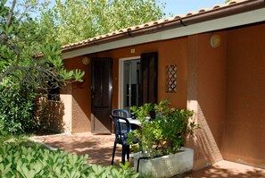 A small, single-story building with a brown exterior and a tiled roof. There is a door with a window and a small table with a chair outside.