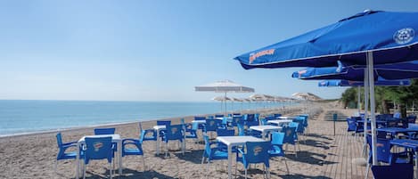 Un restaurante frente a la playa con sillas y sombrillas azules, una terraza de madera y vistas al mar.