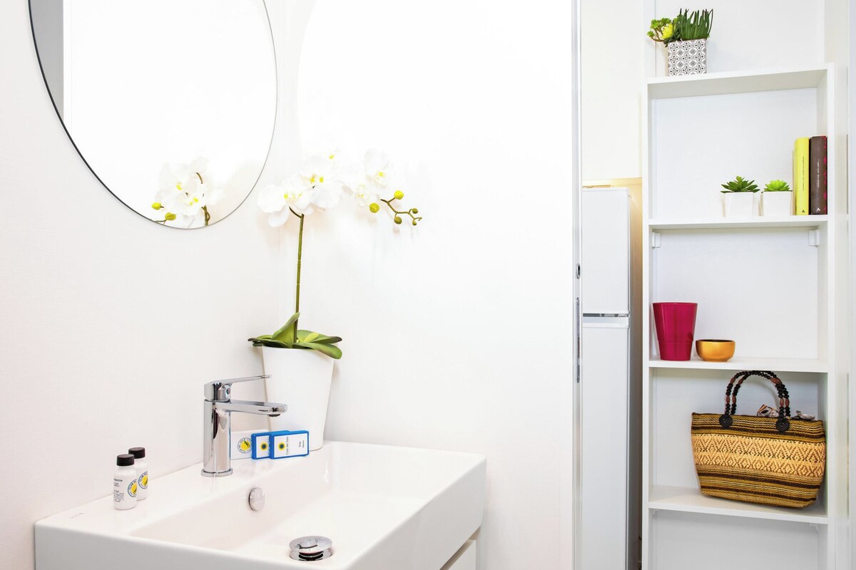 A bathroom with a white sink, mirror, and orchid plant. There is a shelf with books, a red cup, and a woven basket.