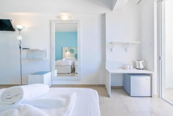 A modern hotel room with a bed, a desk, a small refrigerator, and a mirror reflecting a bathroom with a blue wall and a white sink. - Santa Maria City Hotel (Rhodes)