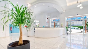 A modern hotel lobby with a reception desk, potted plants, and a view of the street outside. - Santa Maria City Hotel (Rhodes)
