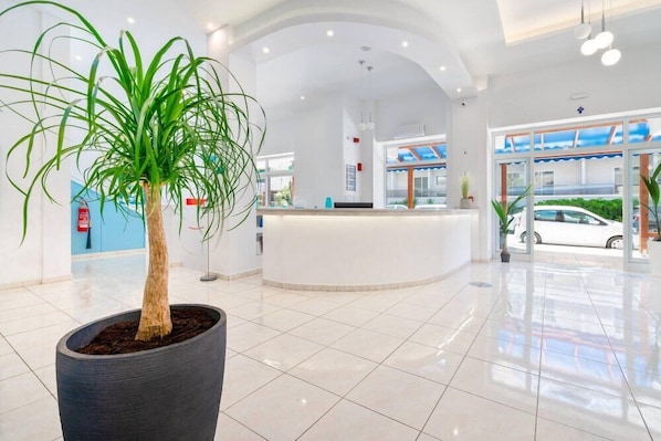 A modern hotel lobby with a reception desk, potted plants, and a view of the street outside. - Santa Maria City Hotel (Rhodes)