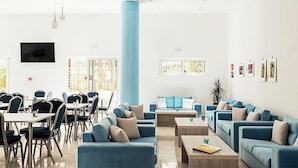 A spacious room with blue sofas, a dining area with tables and chairs, a TV mounted on the wall, and framed pictures on the wall. - Santa Maria City Hotel (Rhodes)