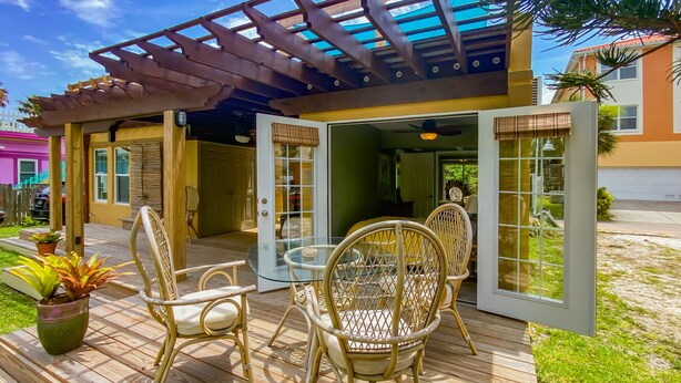 Une terrasse en bois avec des chaises en osier et une table en verre, une pergola avec un toit bleu et une vue sur une maison avec un portail de garage.