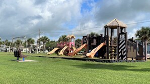 Un parque infantil con toboganes y estructuras para escalar, rodeado de césped y palmeras.