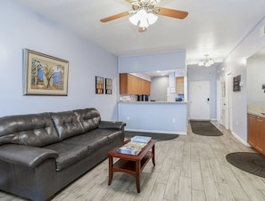 A living room with a leather couch, a coffee table with books, a ceiling fan, and a kitchen in the background. - NEAR DISNEY! 1 BR Condo - SLEEPS 4, Tennis, Baby Pool, Tennis, Shuffleboard, etc (Kissimmee)