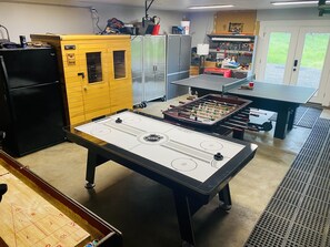 A room with an ice hockey table, a foosball table, and a ping pong table. There are cabinets and a refrigerator in the room.