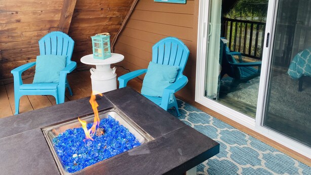 Um deck de madeira com duas cadeiras Adirondack azuis, uma mesa pequena branca e um espaço para fogueira.