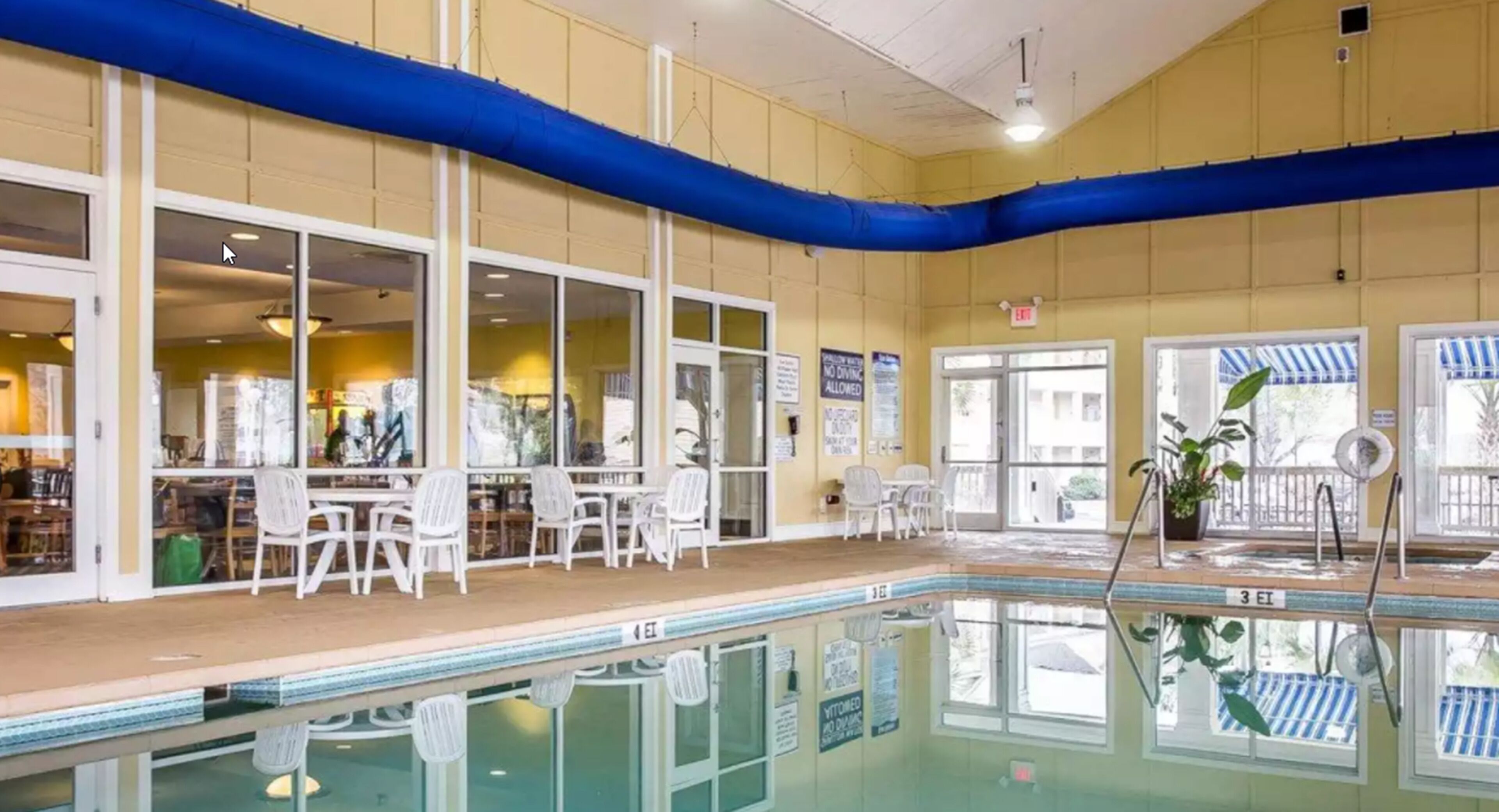 Una piscina interior con agua cristalina, rodeada de sillas y mesas blancas, y amplias ventanas que permiten la entrada de luz natural.