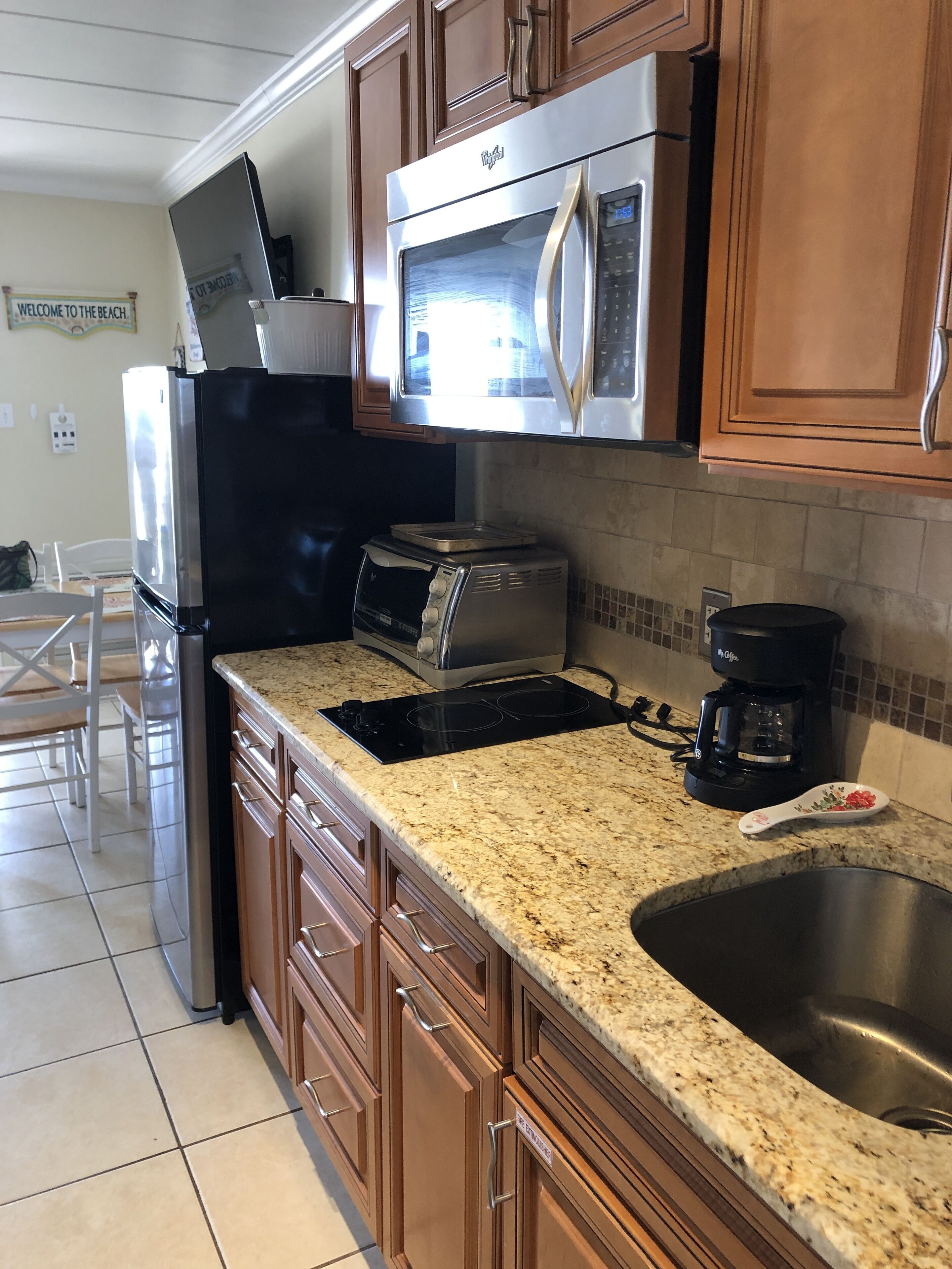 A kitchen with wooden cabinets, a stainless steel refrigerator, a microwave, a toaster, a coffee maker, and a granite countertop.