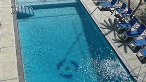 A rectangular swimming pool with blue water, surrounded by lounge chairs and a deck area with tables and umbrellas.