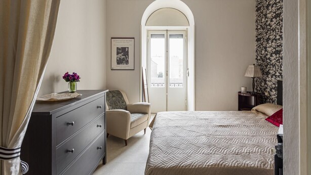 A bedroom with a bed, a dresser, a chair, a nightstand, a lamp, a window with an arch, and a door. - Hostmate - Milano - Cantoni - Washington  (Milano)