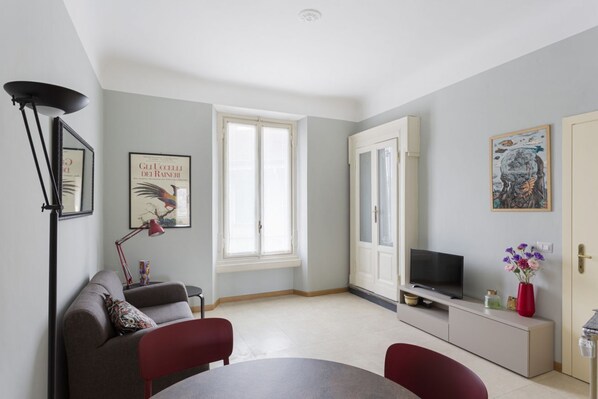 A modern living room with a sofa, a dining table, a TV on a cabinet, a large window, and artwork on the walls.
