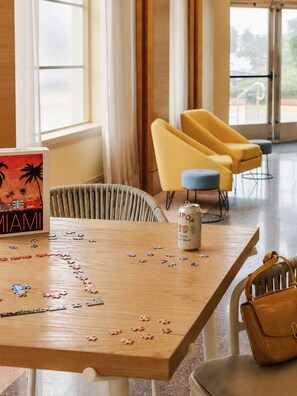 A wooden table with a puzzle, a can of beer, and a book titled 'MIAMI' in a room with yellow chairs and a view through a window. - The Abbey Hotel Miami by M11 Collection (Miami Beach)