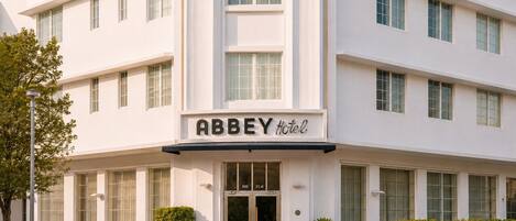 Un edificio blanco con un letrero que dice 'Abbey Hotel'. Se observan varias ventanas en la fachada, una escalera que lleva a la entrada y un pequeño jardín con plantas.