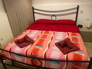 Cama bem arrumada com um cobertor vermelho e duas toalhas dobradas em uma estrutura de metal, em um quarto com porta de madeira e parede branca.