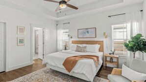 Ein Schlafzimmer mit Bett, Deckenventilator, Nachttisch, Stuhl und einem Fenster mit Vorhängen.