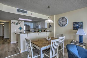 A dining area with a wooden table and white chairs, a kitchen in the background, a clock, and a decorative wall piece. - Bay Watch Condo w/ Oceanfront Balcony & Beach View (North Myrtle Beach)