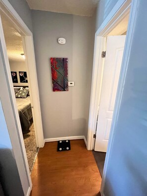 A hallway with a gray wall, a colorful abstract painting, a thermostat, and a black object on the floor.