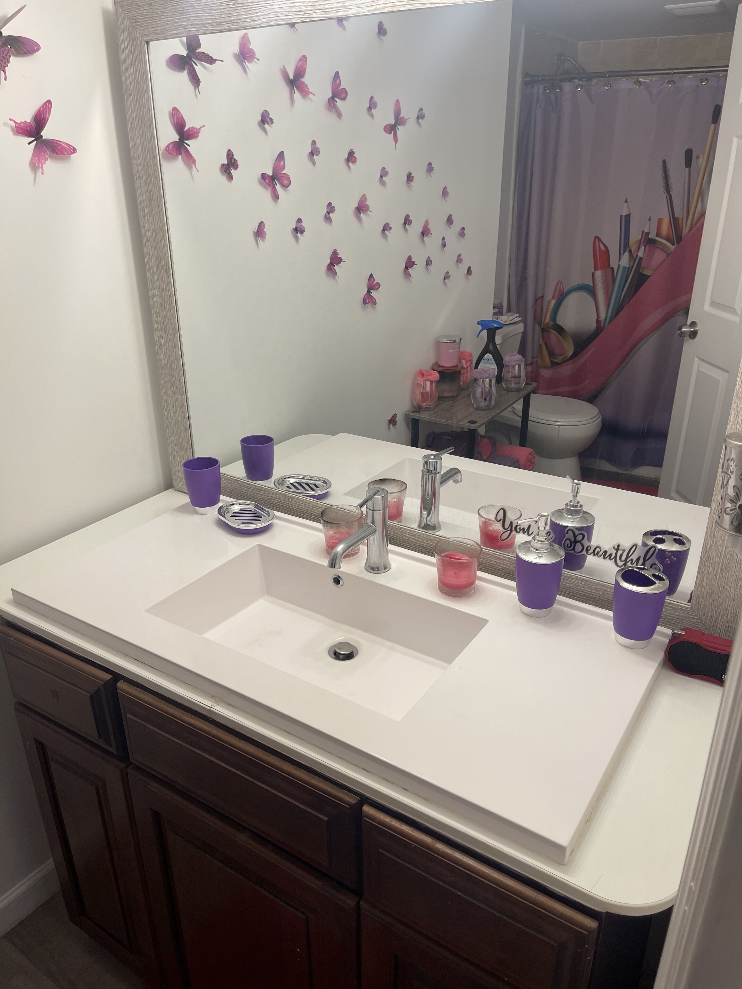 A bathroom with a white sink, purple cups, and a mirror reflecting a toilet and shower curtain.