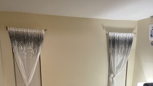 A room with a bed covered in a leopard print bedspread, two windows with sheer curtains, a bedside table with toiletries, and a power outlet on the wall. - GORGEOUS 1/1 CONDO (Lauderdale Lakes)