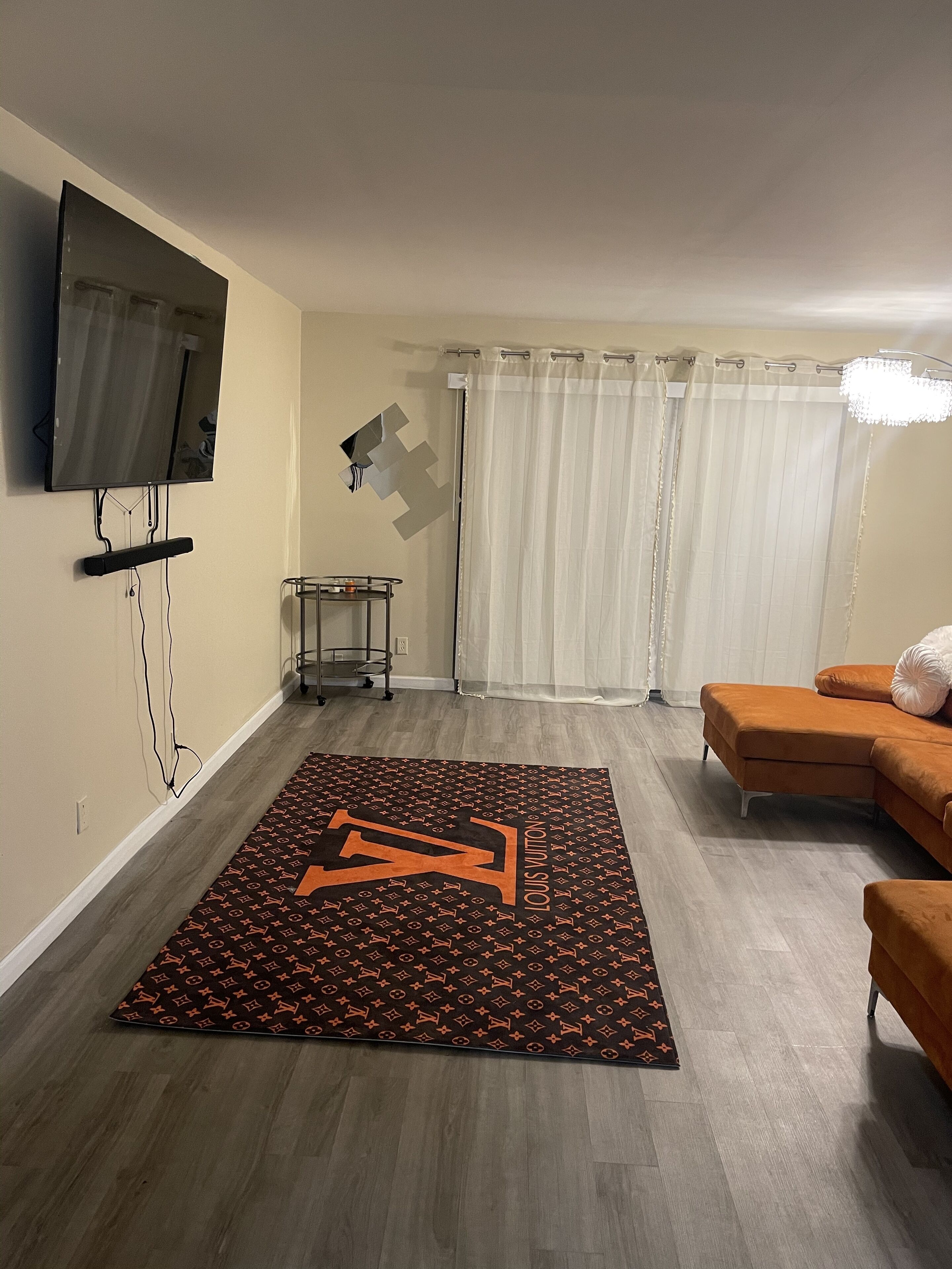 A living room with a flat-screen TV mounted on the wall, a Louis Vuitton patterned rug, an orange sofa, and a white curtain.