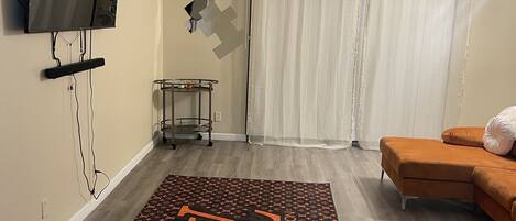 A living room with a flat-screen TV mounted on the wall, a Louis Vuitton patterned rug, an orange sofa, and a white curtain.