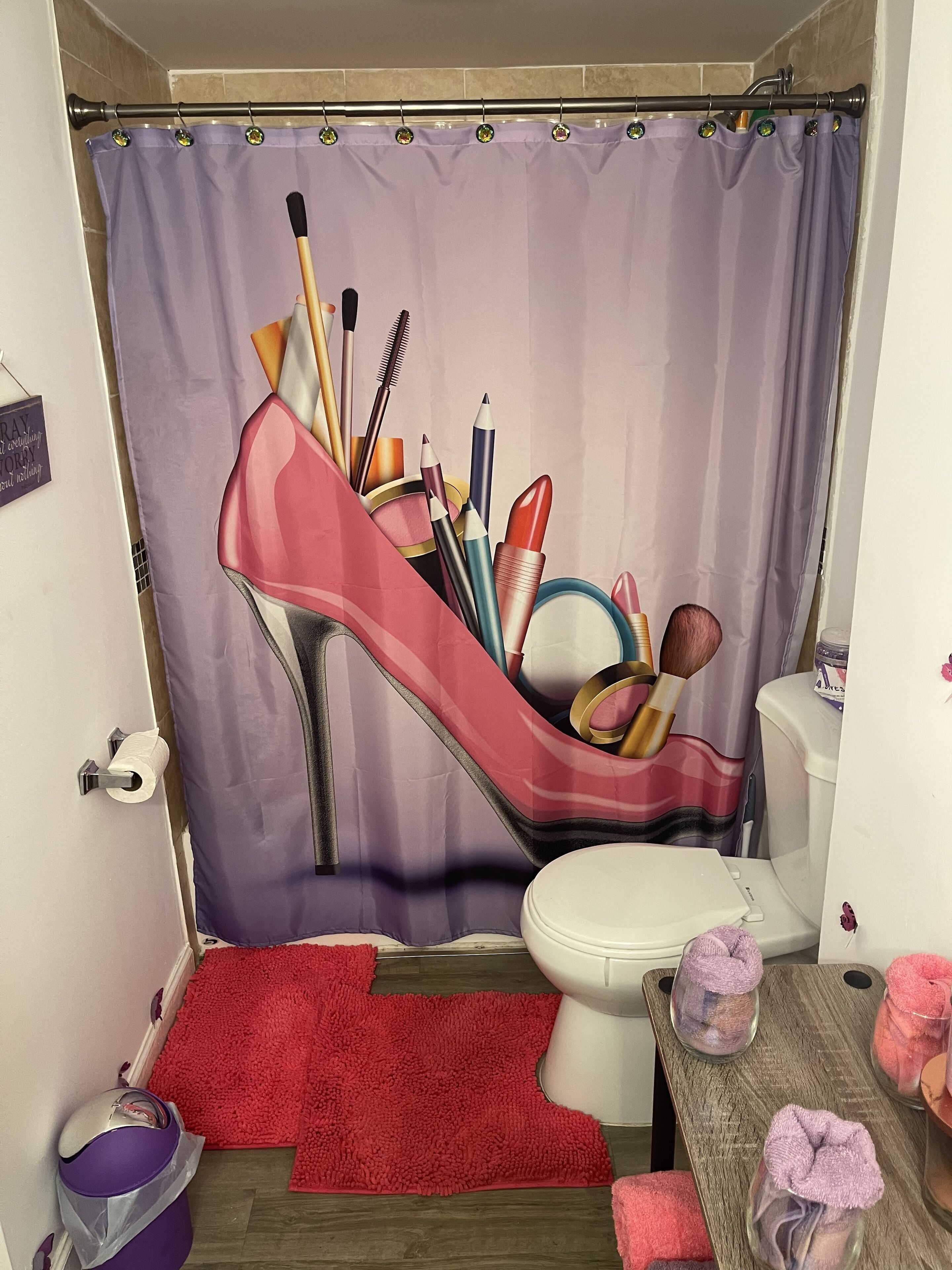 A bathroom with a shower curtain depicting a high-heeled shoe, a toilet, a red bath mat, a trash bin, and a wooden bench with towels.