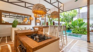 A kitchen with a wooden island, hanging lights, and a view of a pool and greenery outside. - Private Pool Courtyard | Three Suites | Cafe Walk (Kerobokan)