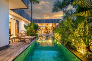 A modern outdoor pool area with lounge chairs, a wooden deck, and lush tropical plants.